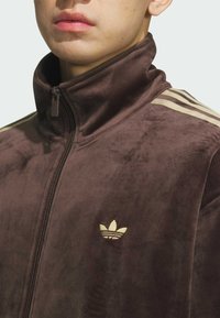 Brown velour zip-up jacket featuring a high collar, gold three-stripe accents on shoulders, and a small gold embroidered logo on the chest.