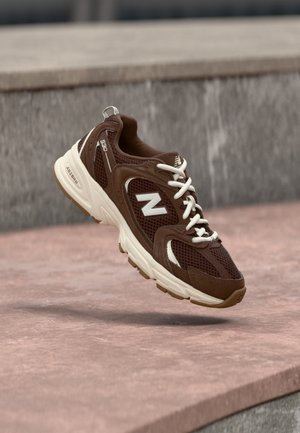 Hnedo-biela teniska New Balance vznášajúca sa nad ružovým kamenným povrchom so sivým betónovým pozadím.