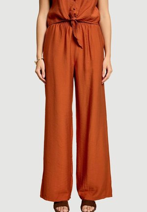 Femme portant un pantalon ample couleur rouille et un haut noué assorti, pieds nus dans des chaussons ouverts marron foncé, debout sur un fond neutre.