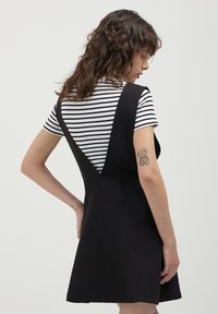 Robe noire sans manches avec un corsage ajusté, une jupe évasée et un dos en V profond, portée sur une chemise à rayures à manches courtes.