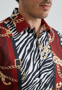 Chemise à manches courtes arborant un motif rayé zèbre rouge et noir, des chaînes dorées et des détails ornés, avec un design de col contrastant.