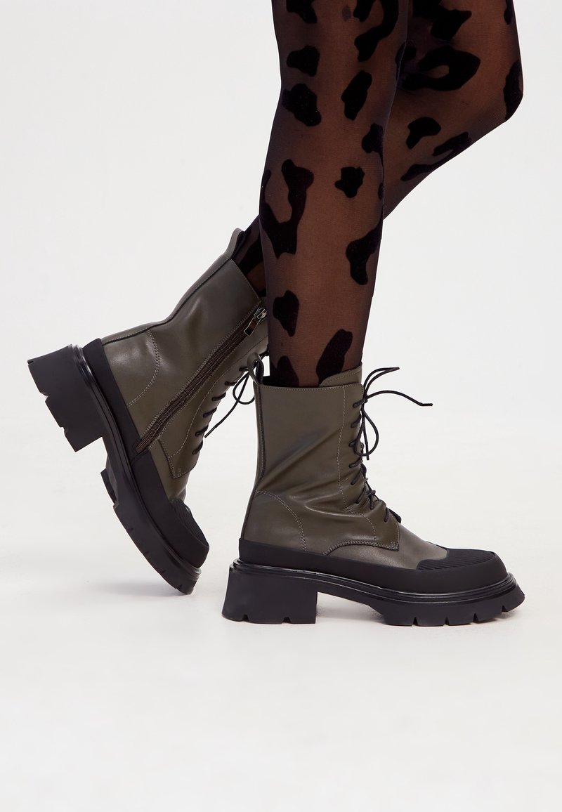 Bottes de combat à lacets couleur olive avec semelles noires épaisses portées avec des collants transparents noirs à motifs.