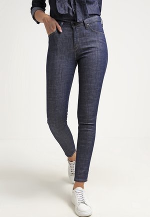 Kvinde iført mørkeblå skinny jeans og hvide sneakers, med hånden i lommen, stående mod en hvid baggrund.