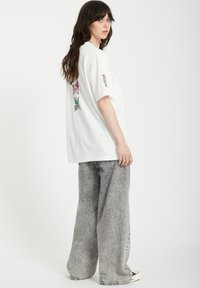 Camiseta blanca de gran tamaño con bordado floral colorido en la parte posterior y en las mangas, combinada con pantalones de mezclilla gris de pierna ancha con un acabado texturizado.
