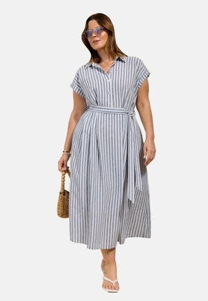 Kvinde iført en blå og hvid vertikalt striped midi kjole med korte ærmer, bælte, solbriller, hvide sandaler og holder en vævet håndtaske.