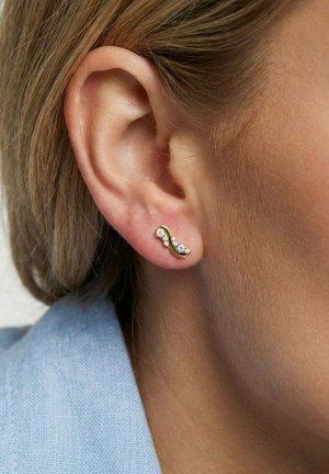 Nærbillede af et øre med en guldbueformet ørestik med diamanter, båret af en person med lysebrunt hår og lys hud.