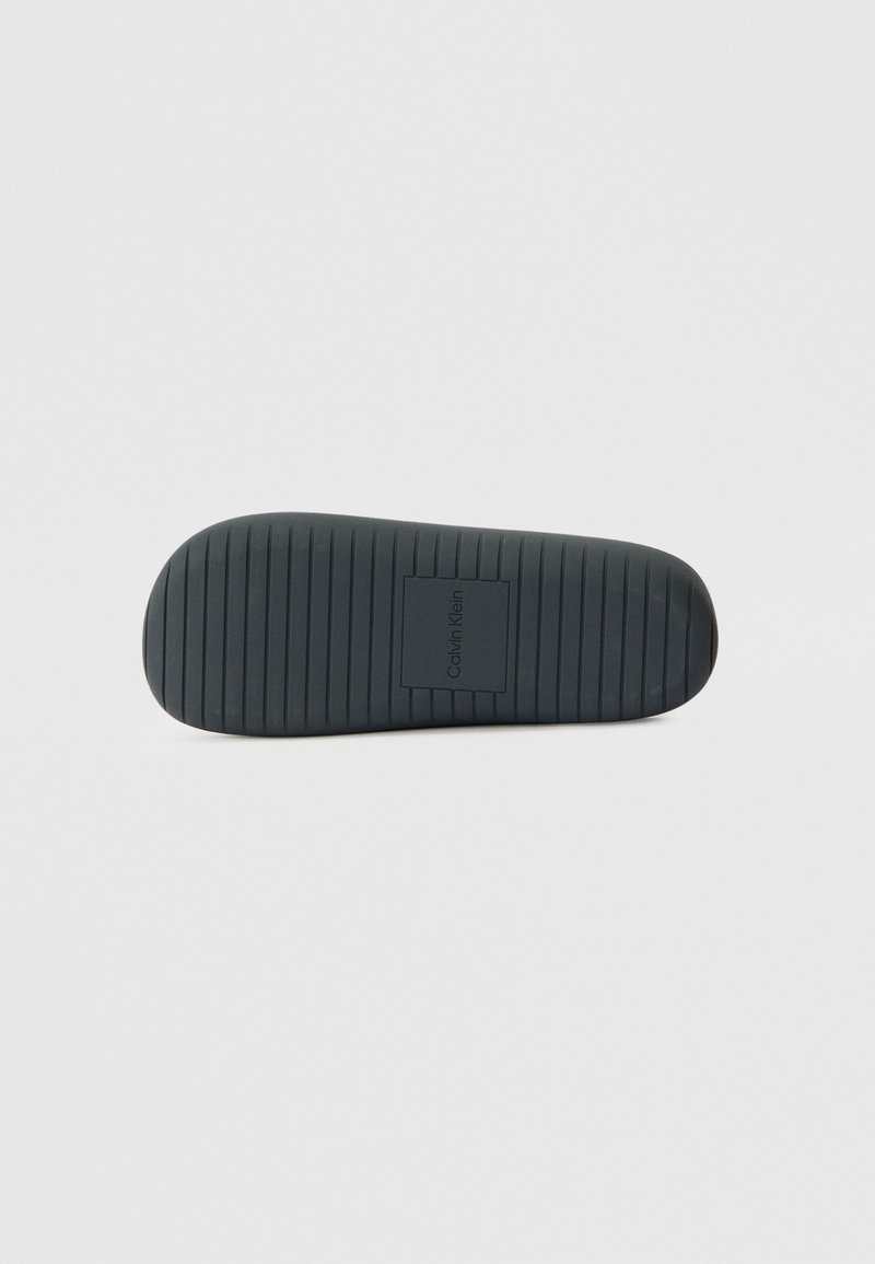 Schwarze Gummisohle mit horizontalen Rillen, abgerundetem Zehenbereich und eingeprägtem Logo in der Mitte. Glatte Textur mit strapazierfähiger Oberfläche.