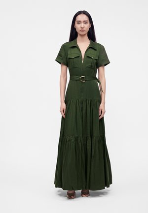 Femme debout portant une longue robe vert foncé à manches courtes, fermeture éclair devant, poches poitrine et taille ceinturée avec une boucle dorée.