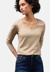 Beige geripptes Langarmoberteil mit V-Ausschnitt. Verfügt über schmale Ärmel und eine taillierte Passform, kombiniert mit dunklen Denim-Jeans.