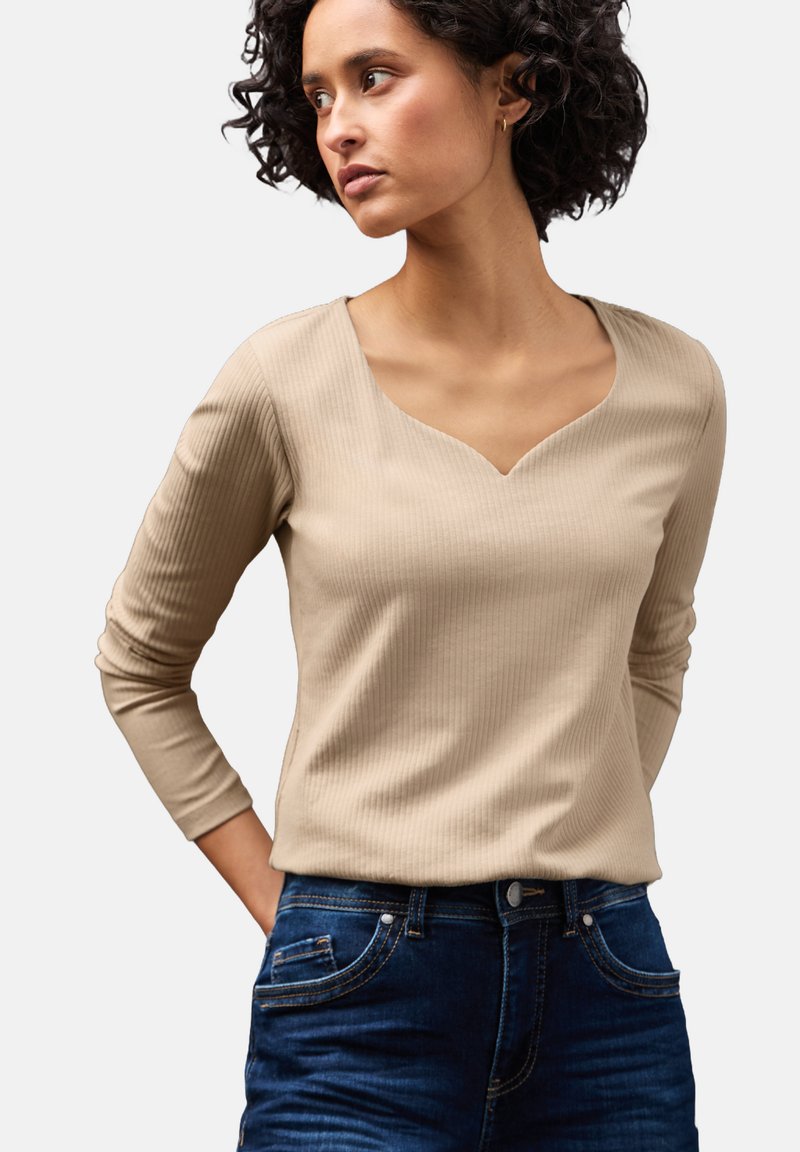 Beige geripptes Langarmoberteil mit V-Ausschnitt. Verfügt über schmale Ärmel und eine taillierte Passform, kombiniert mit dunklen Denim-Jeans.