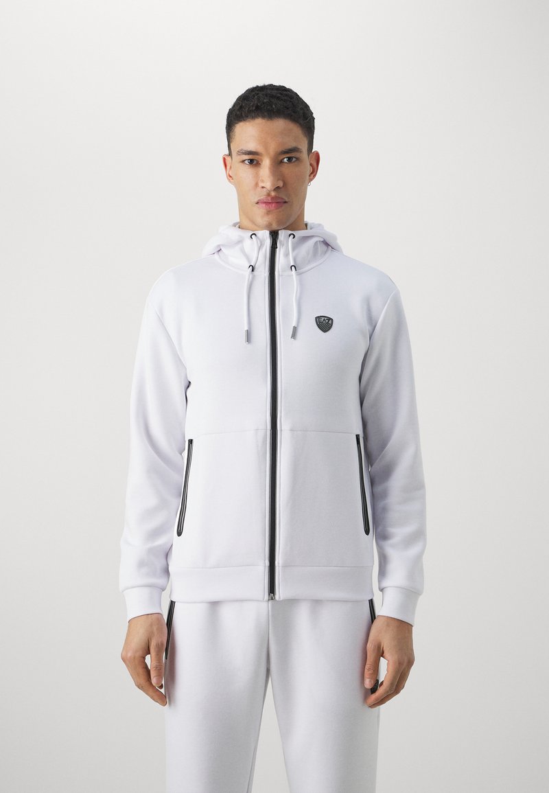 Felpa bianca con zip e cappuccio con coulisse, dettagli neri sulla zip, tasche laterali e un logo sul petto, abbinata a pantaloni coordinati.