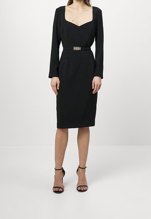 Vrouw draagt een zwart knielang jurkje met lange mouwen, een vierkante halslijn, een tailleband en zwarte hoge hakken met enkelbandje tegen een effen achtergrond.