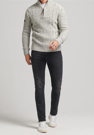 Homme portant un pull à col montant zippé gris clair en maille torsadée, jean noir slim, baskets blanches et une bague argentée à la main droite.