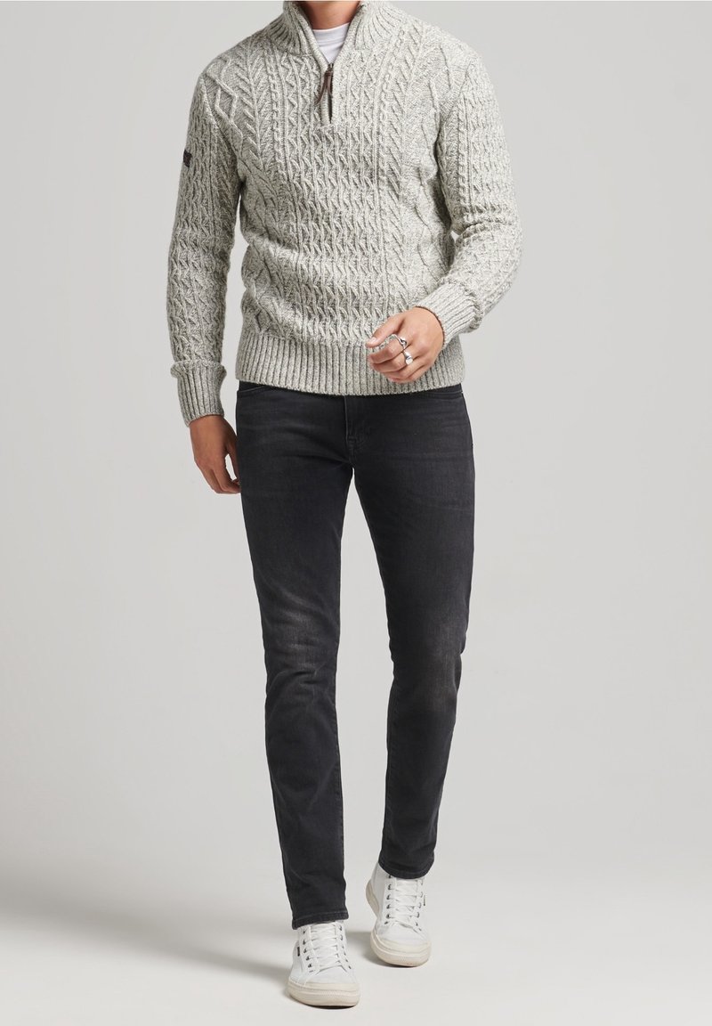 Homme portant un pull à col montant zippé gris clair en maille torsadée, jean noir slim, baskets blanches et une bague argentée à la main droite.