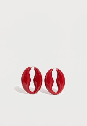 Par rdečih, valovitih, sijajnih uhanov obročkov s srebrnimi kovinskimi zaponkami na belem ozadju.