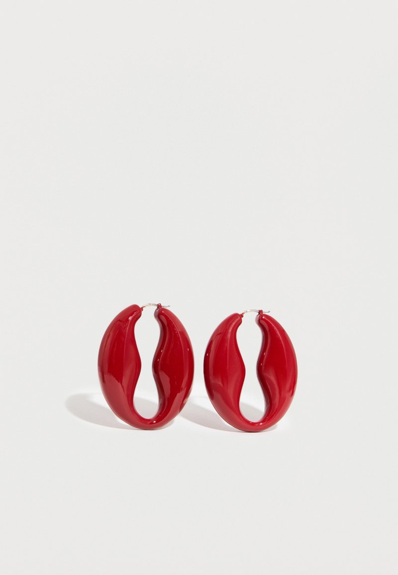 Ζεύγος γυαλιστερών, κόκκινων, κυματιστών κρίκων σκουλαρικιών με ασημένιες μεταλλικές αγκράφες σε λευκό φόντο.