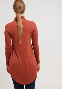 Långärmad, rostfärgad topp med hög krage och en böjd sidsöm, tillverkad av ett mjukt, stretchigt material, åtsittande vid armarna.