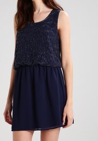 Marindress har en pärlbroderad topp med vågmönster och en flödande kjol. Ärmlös design, slätt tyg och rynkad midjedetalj.