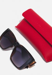 Occhiali da sole oversize neri e tartarugati con lenti sfumate accanto a un astuccio in pelle rossa impresso con il nome del marchio.