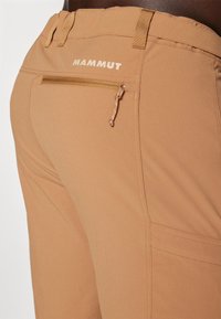Beiga vandringsbyxor med en ficka på ryggen med dragkedja och logotypdetalj. Materialet är strukturerat, med sömmar synliga längs sömmarna.