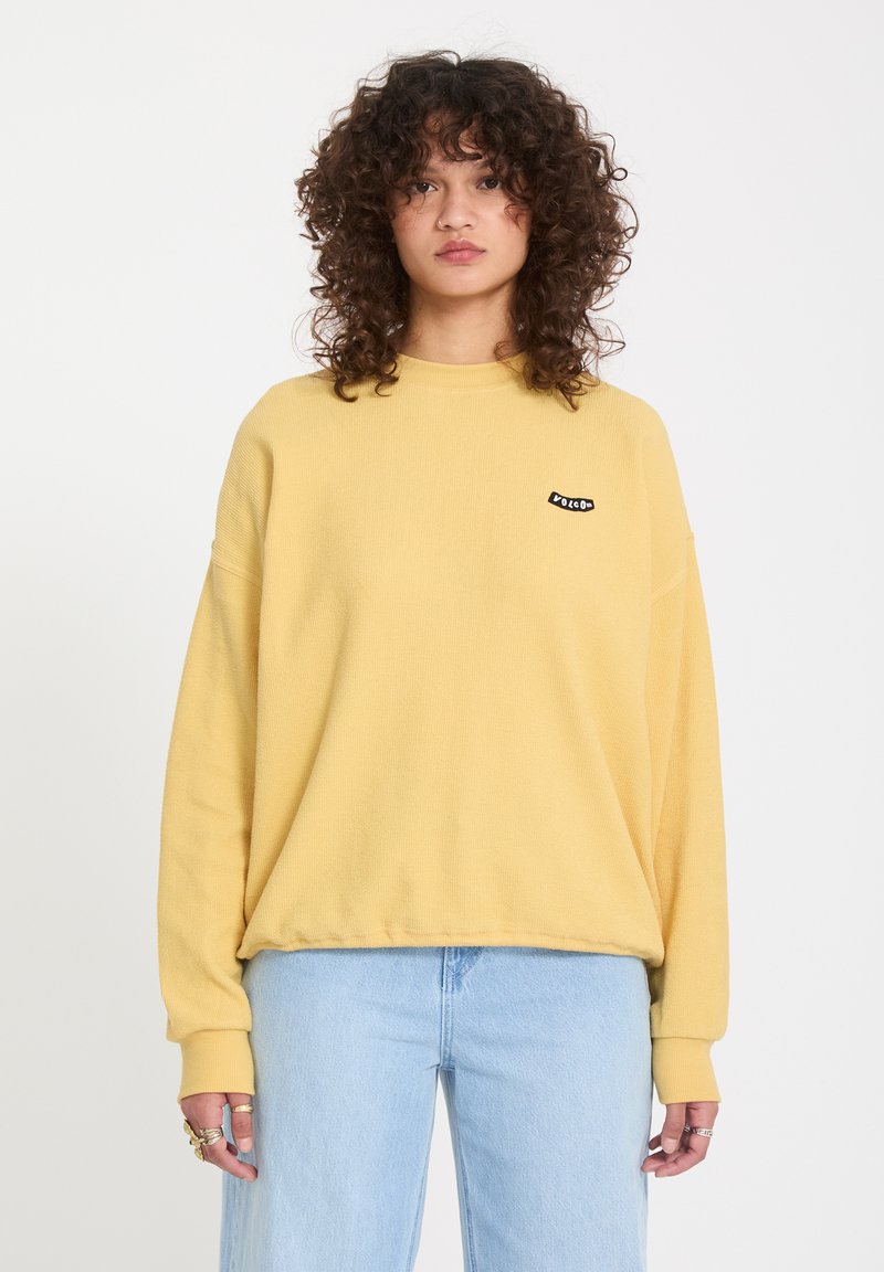 Personne aux cheveux bouclés portant un sweat-shirt jaune ample avec un petit logo et un jean bleu clair, debout devant un fond blanc.