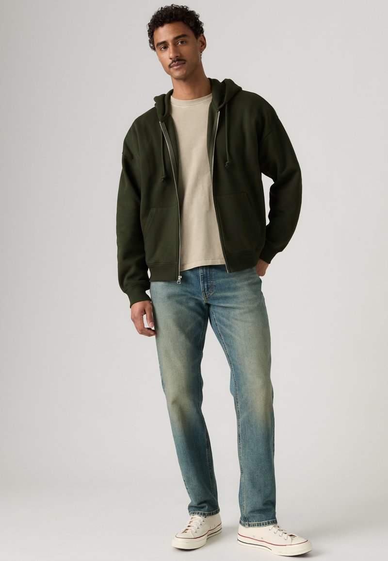 Mand stående iført en mørkegrøn hættetrøje med lynlås, beige t-shirt, blå jeans og hvide sneakers mod en ensfarvet lyse baggrund.