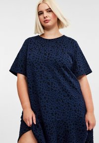 Frau mit blonden Bob-Haaren, die ein lockeres, kurzärmliges dunkelblaues Kleid mit einem dezenten, schwarzen Leopardenmuster trägt und den Saum leicht hält.