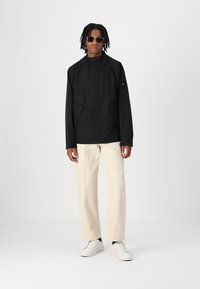 Schwarze wasserdichte Jacke mit Kapuze, Fronttaschen und Reißverschluss. Getragen über hellen Hosen und kombiniert mit weißen Sneakers.