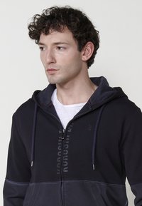 Crna zip-up hoodie sa sivim kapuljačom, koja ima dizajn logotipa i teksta sprijeda. Materijal dukserice s kontrastnim panelima.