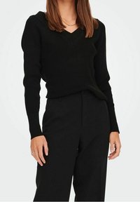 Mujer vistiendo un suéter negro de canalé con escote en V y pantalones negros de cintura alta frente a un fondo blanco liso.