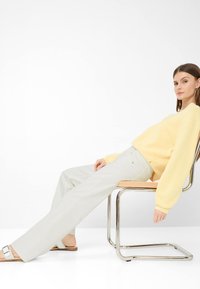 Donna sdraiata su una sedia con struttura in metallo, indossa un maglione giallo chiaro, pantaloni bianchi e sandali bianchi a ciabatta su sfondo bianco.