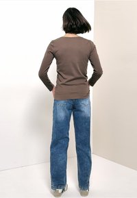 Hnědý top s dlouhým rukávem, přiléhavý střih, v kombinaci se světle modrými džínami s rovnými nohavicemi. Boty jsou béžové se zelenými akcenty. Pozadí je neutrální.