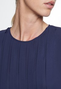 Abito blu navy con collo tondo e pieghe verticali. Il tessuto sembra leggero con una consistenza liscia. Nessun hardware visibile.