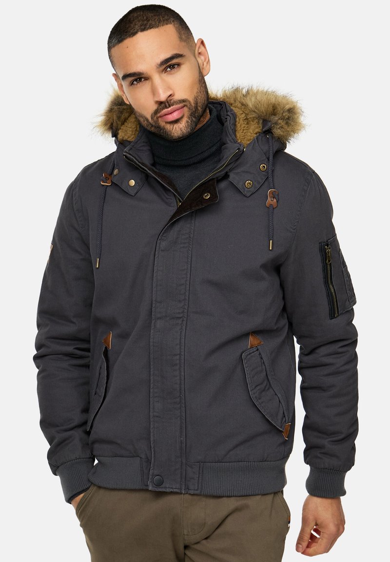 Chaqueta gris con capucha forrada de piel sintética, mangas con cremalleras y acentos marrones contrastantes. Tiene un cierre frontal con botones y múltiples bolsillos.