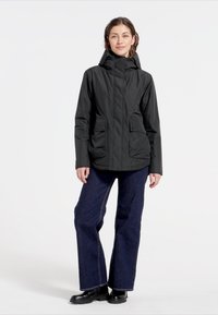 Svart vattentät jacka med huva, framdragkedja och två fickor med klaff. Bärs med vida blå jeans och svarta stövlar. Slät textur.