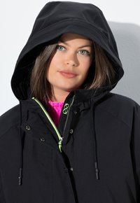 Černá vodotěsná bunda s kapucí, vybavená jasně zeleným zipem a stahovací šňůrkou, a s texturovanou vnitřní vrstvou, která je viditelná pod povrchem.