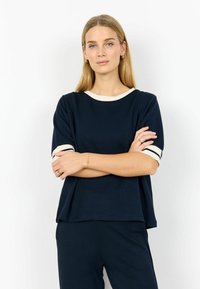 Temno modra majica s kratkimi rokavi, obrobljena s kremnim trakom in črtastimi poudarki na rokavih. Ohlapen kroj, mehka tekstura, primerna za vsakodnevno nosenje.