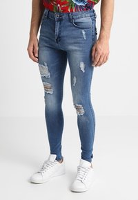 Eng anliegende blaue Jeans mit Rissdetails, mittelhohem Bund und Fünf-Taschen-Design, kombiniert mit weißen Turnschuhen.