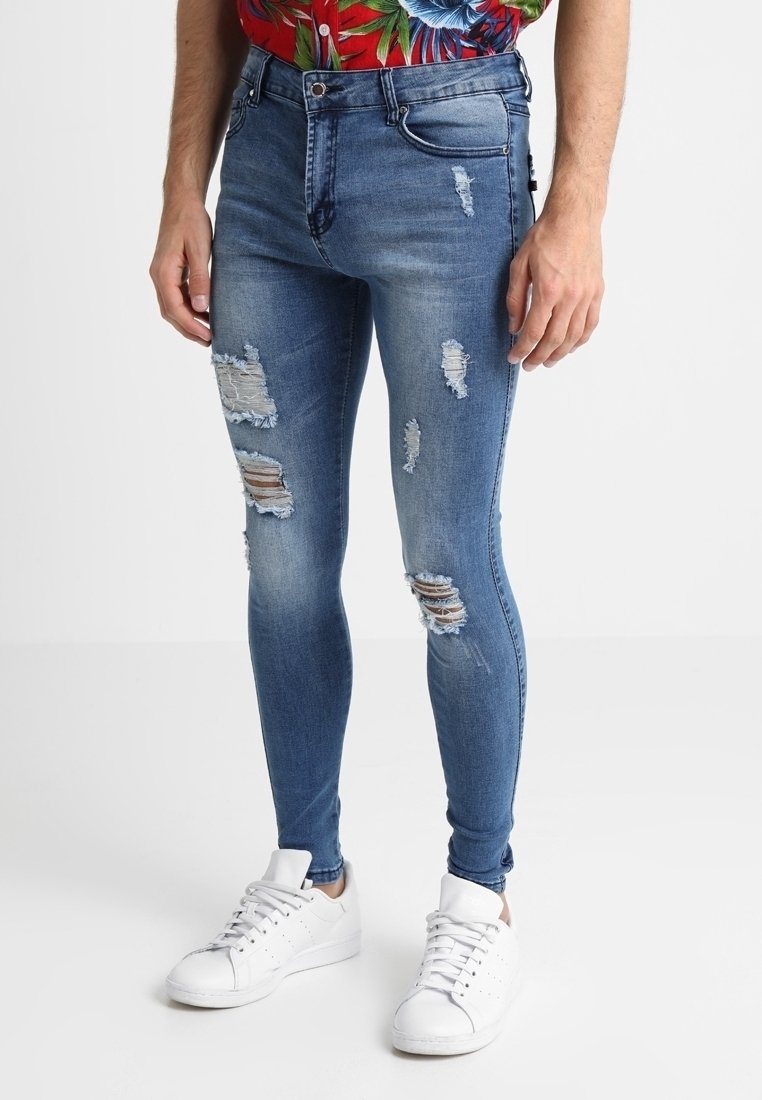 Eng anliegende blaue Jeans mit Rissdetails, mittelhohem Bund und Fünf-Taschen-Design, kombiniert mit weißen Turnschuhen.