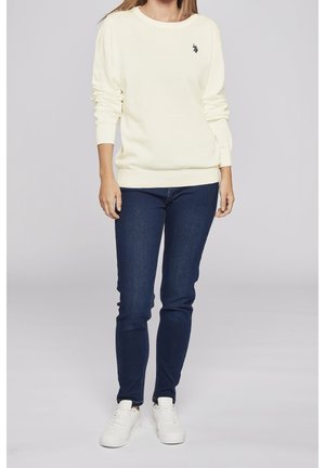 Sweat-shirt en maille jaune clair avec encolure ronde, poignets et ourlet côtelés, associé à un jean bleu foncé et des baskets blanches. Détail du logo noir visible.