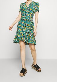 Robe portefeuille à fleurs en vert, avec des manches courtes bouffantes, un col en V et un ourlet fringant. Associée à des sandales à plateforme noires.