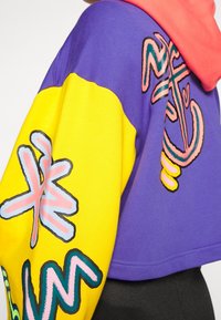 Hoodie cropped con sezioni viola e gialle, con grafiche rosa e nere. Texture liscia e contrasti di colore vivaci.