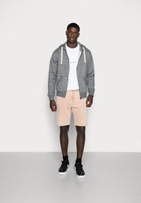 Grijze rits hoodie met trekkoorden, witte grafische T-shirt, lichtroze shorts en zwarte sneakers met witte zolen. Effen achtergrond.