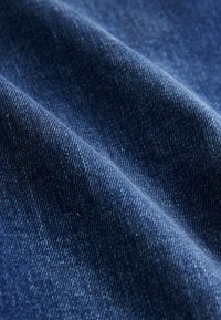 Gros plan sur un tissu en denim bleu texturé avec des motifs de tissage visibles et des plis doux créant des ombres.