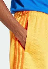 Yellow mesh shorts with orange stripe accents, elastic waistband, and side pockets. Close-up view shows texture and fit against blue shirt.