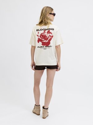 Femme portant des lunettes de soleil, un T-shirt oversize crème avec une rose rouge et le texte "BLOOMING", un short noir et des bottines beiges, debout sur un fond blanc.