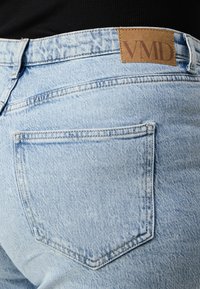 Back pocket and waistband of light blue denim jeans with stitched seams and a tan leather patch labeled "VMD" worn with a black top.