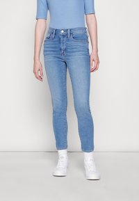Jeans azul claro de cintura alta, ajuste slim e detalhes ligeiramente desgastados. Usado com ténis brancos e meias até ao tornozelo.