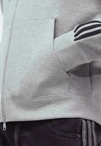 Sivá mikina so zipsom, so predným vreckom a tromi čiernymi pruhmi na rukáve. Mäkka textúra a voľný strih.