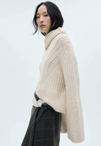 Maglione a coste beige con colletto alto, dotato di maniche ampie e spacco laterale. Una cintura nera con fibbia metallica mette in risalto la vita.